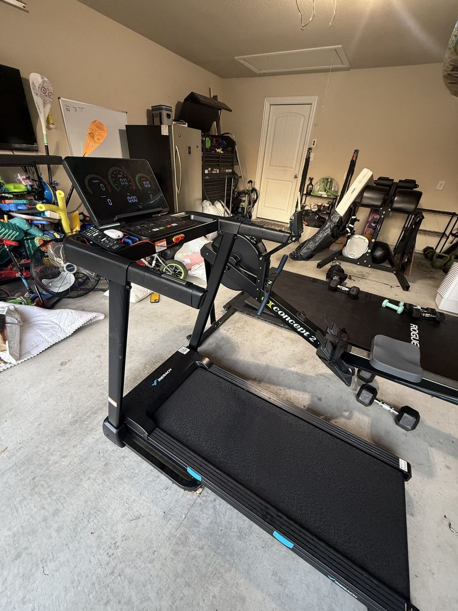 My MERACH T12 Pro treadmill set up in the garage beside a rower and a rack of dumbbells, console lit and ready